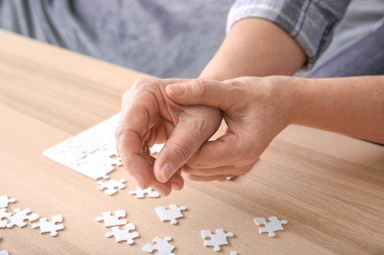 Close-up of an elderly person's hands, one gripping the other, showing signs of stiffness or tremors while working on a jigsaw puzzleâ€”symbolizing the effects of Parkinsonâ€™s disease on fine motor skills and daily activities